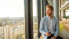 Ein lächelnder Mann im Blazer sitzt mit einem Smartphone am Fenster eines modernen Büros und blickt auf die Stadt.