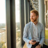 Ein lächelnder Mann im Blazer sitzt mit einem Smartphone am Fenster eines modernen Büros und blickt auf die Stadt.