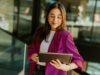 Eine Frau im lila Blazer blickt lächelnd auf ihr Tablet in einem modernen Büro.