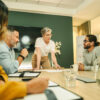 Vier Kollegen bei einer Teambesprechung in einem modernen Büro; eine Frau steht am Tisch und erklärt Dokumente.
