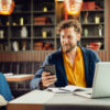 Mann in einem Café am Laptop mit Smartphone in der Hand