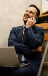 Makler der mit Laptop auf dem Schoß lachend am Smartphone telefoniert