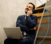 Makler der mit Laptop auf dem Schoß lachend am Smartphone telefoniert